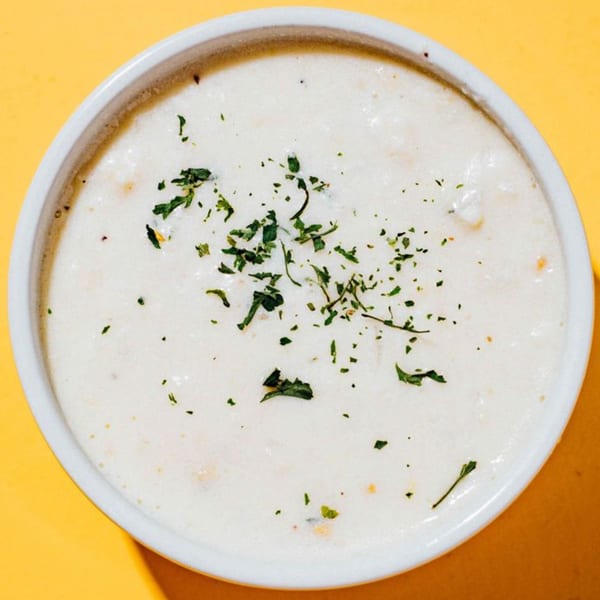 A white bowl of creamy clam chowder, garnished with fresh parsley, on a yellow background.