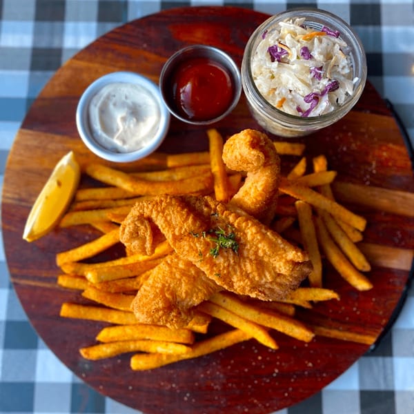 Golden fried fish and chips with coleslaw, tartar sauce, ketchup, and a lemon wedge on a wooden board.